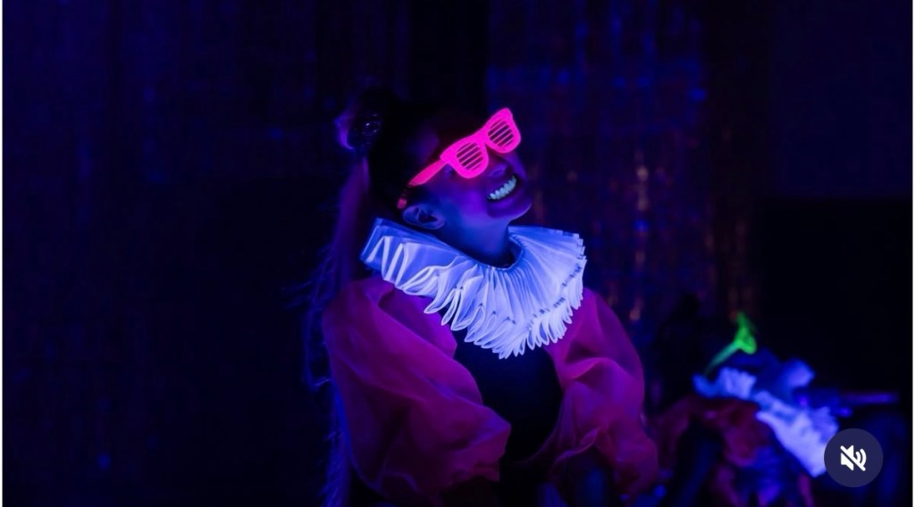 A girl with pink sunshades glows as she sings on stage under a black light