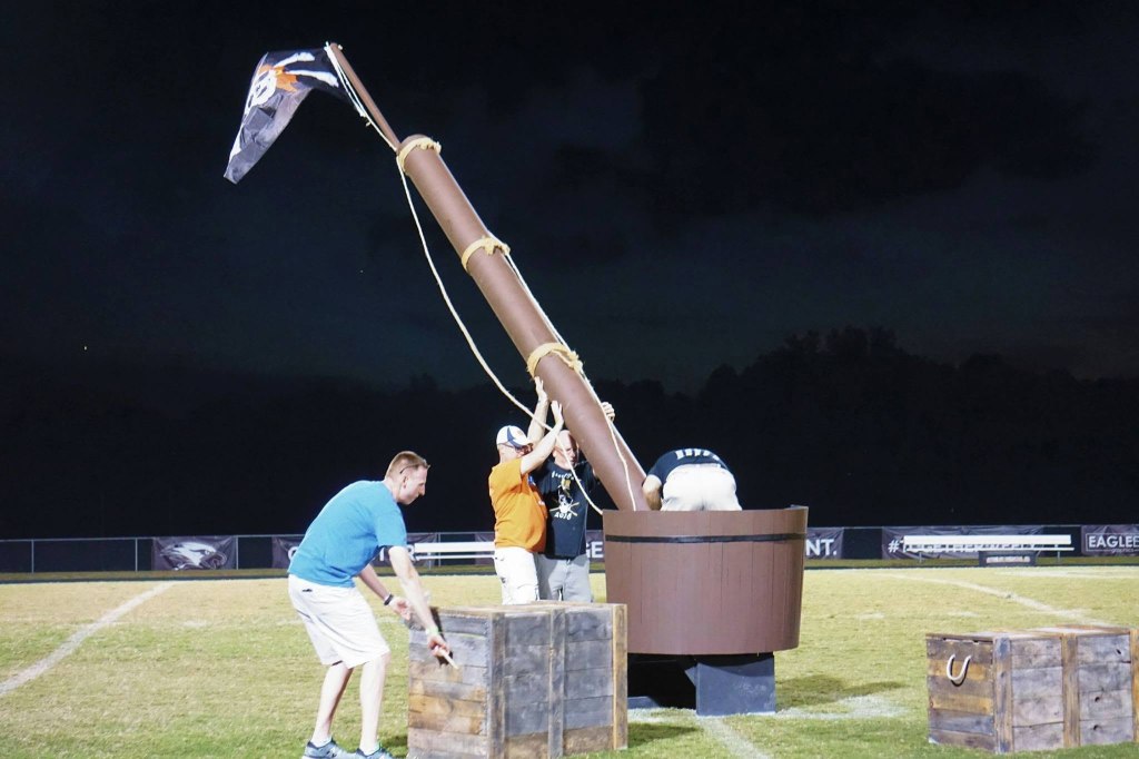 Four men put shipping trunks and raise a pirate ship mast on a football field.