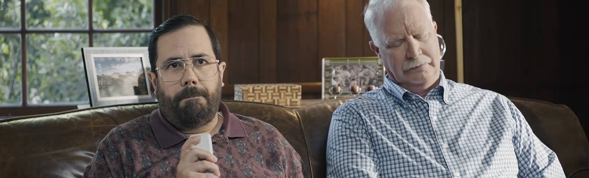 Two men sitting on a couch with one holding a TV remote