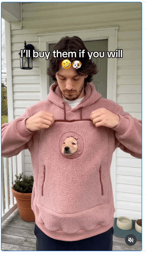 A man wearing a hoodie with a puppy sticking his head through a hole in the center.