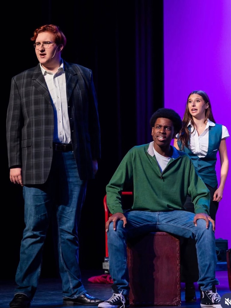 A young man in a blazer singing on stage with another young man in a green jacket and a young woman dressed as a flight attendant.